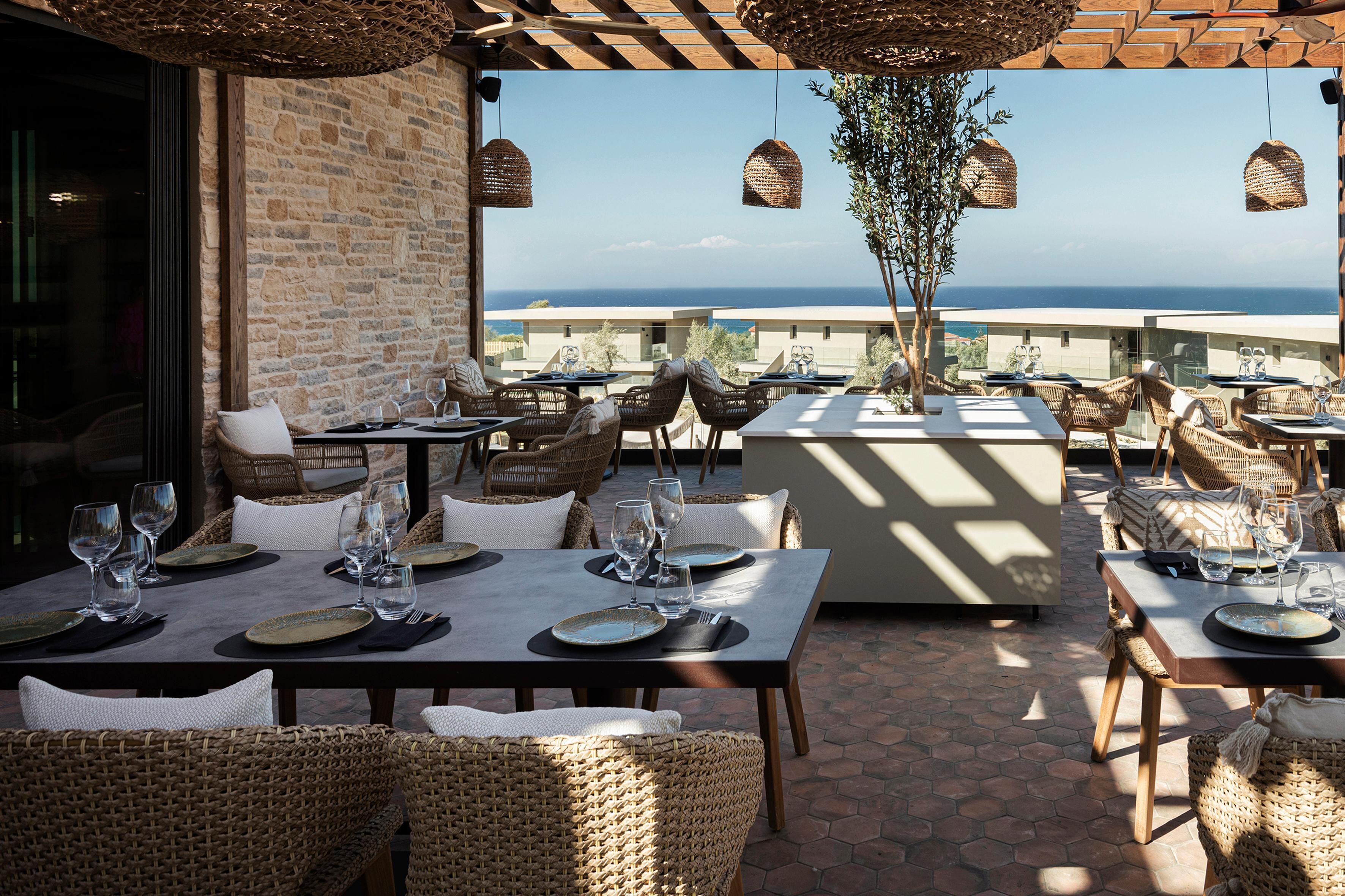 An outdoor dining area with wicker furniture and tables set for dining, overlooking a scenic view of the ocean.