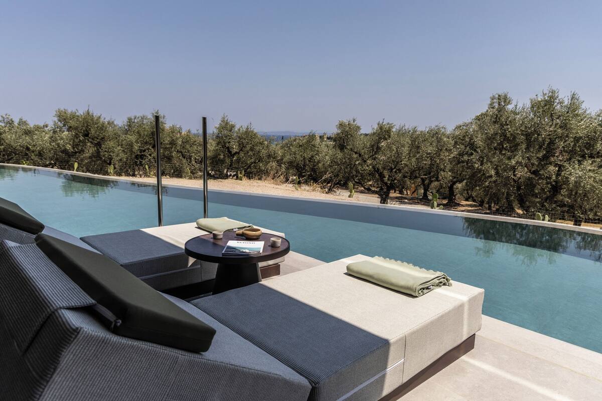 A poolside area with a sofa and a small table, overlooking a clear pool and a landscape of trees.