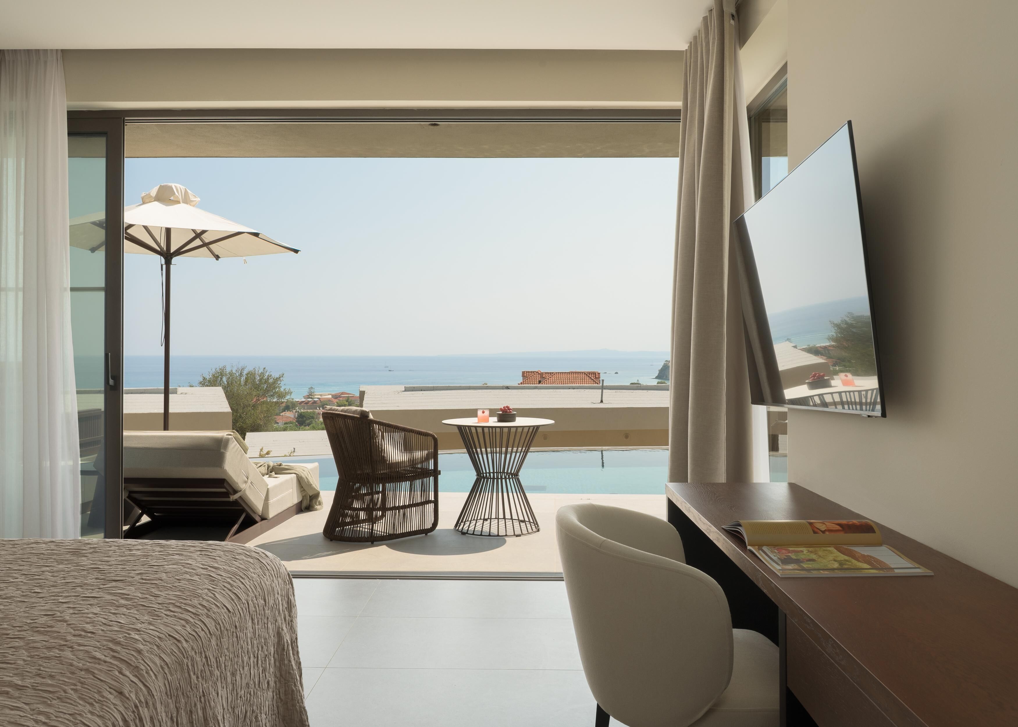 Habitación de hotel con balcón, cama, escritorio, silla, mesita y un amplio ventanal con vista al mar.