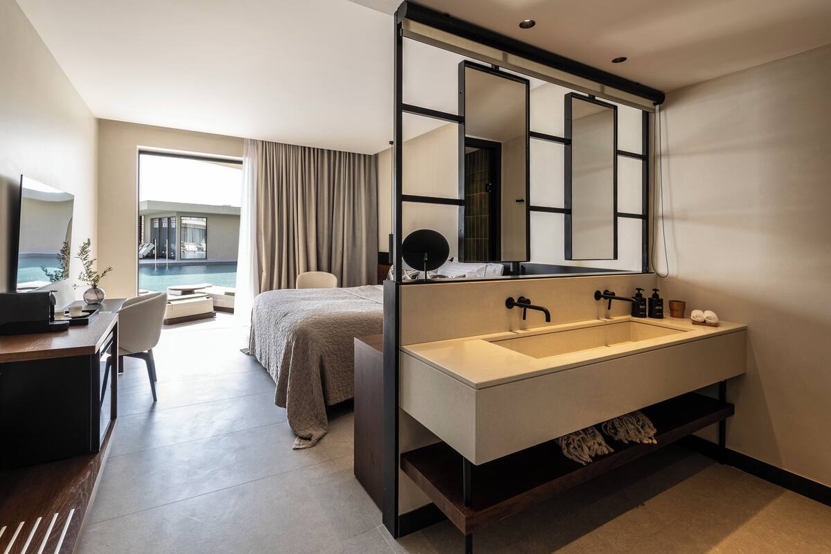A modern hotel room with a large bed, a desk, a large bathroom vanity with a double sink, and a large mirror.