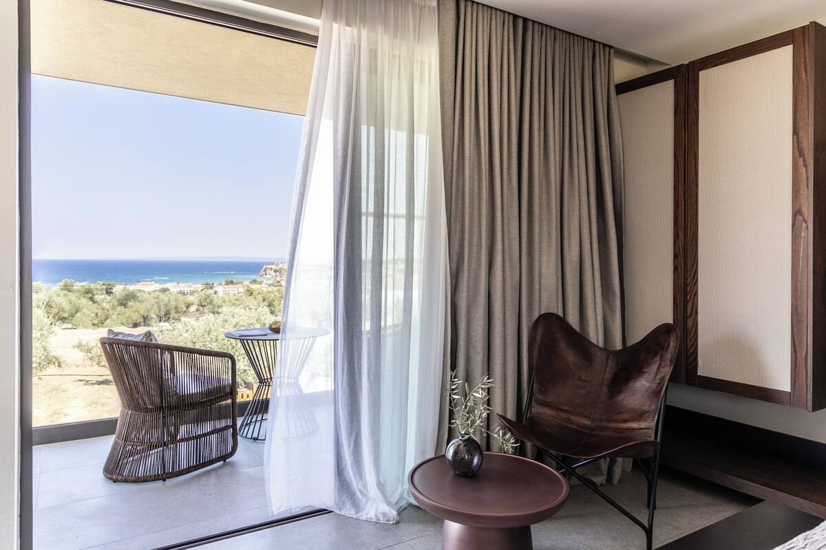 A modern room with a view of the sea, a large window with sheer curtains, a brown leather chair, a small round table, and a wicker chair.