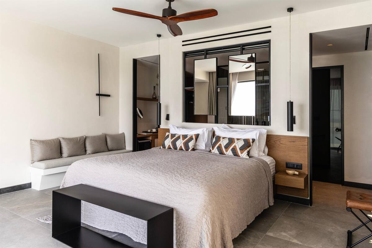 A modern bedroom with a large bed, a ceiling fan, a built-in bench, and a large mirror.
