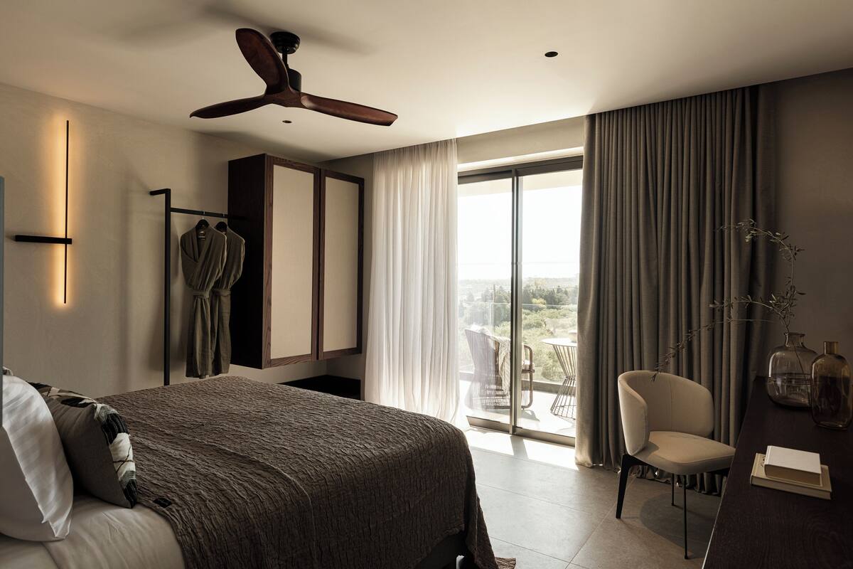 A modern hotel room with a large bed, a ceiling fan, a balcony with a chair, and a view of the outdoors.
