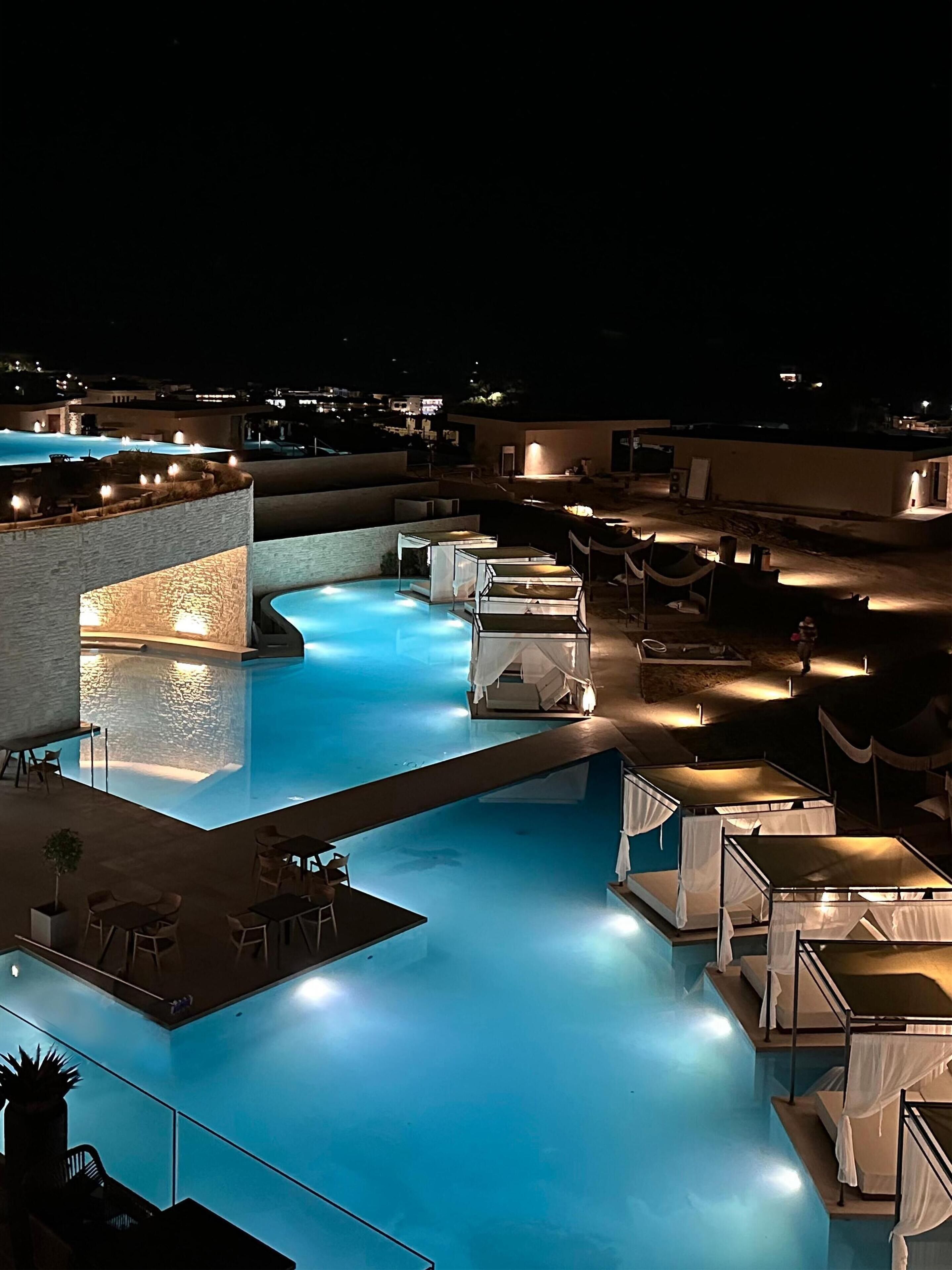 A nighttime view of a swimming pool area with multiple lounge chairs and umbrellas, surrounded by buildings.