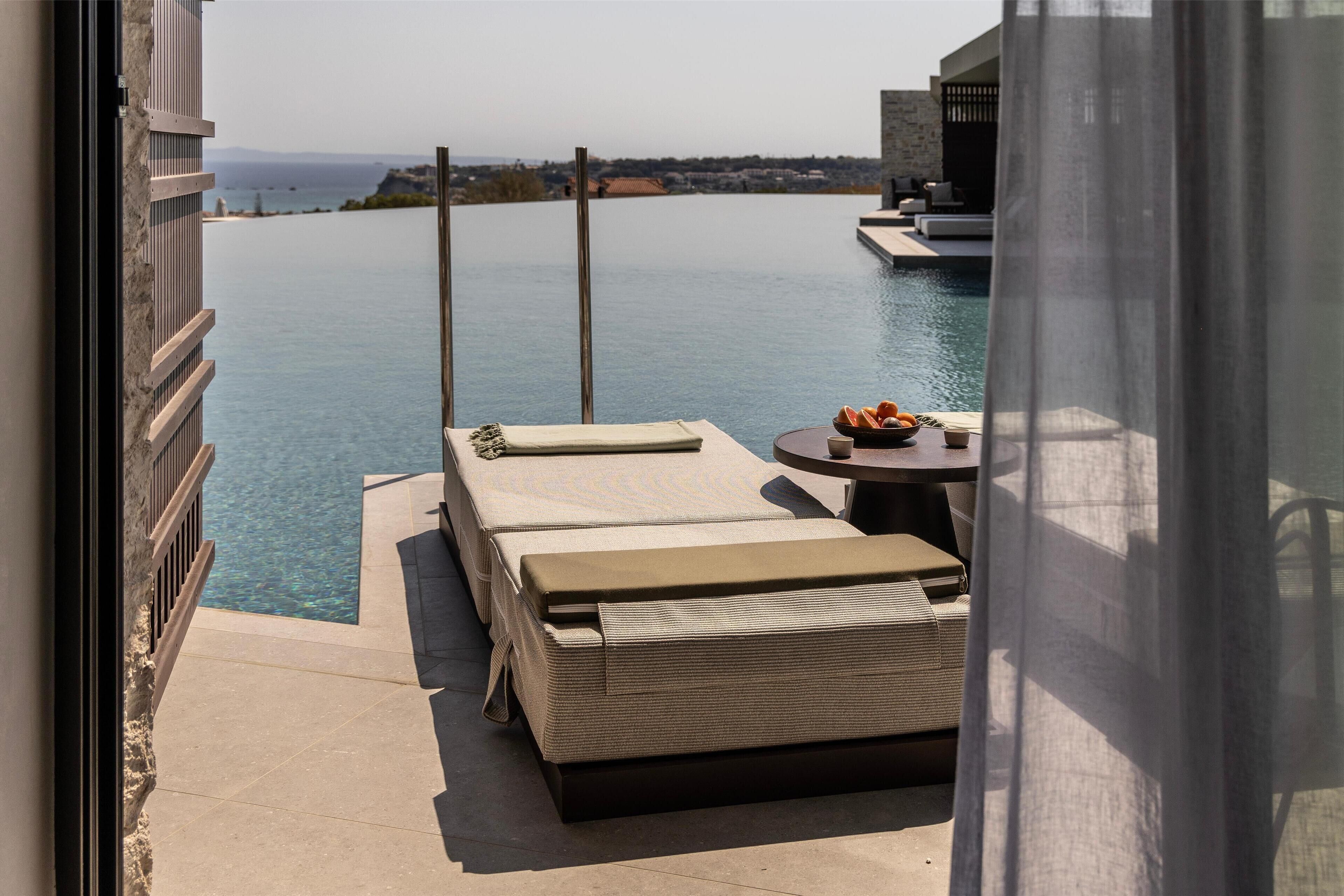 A balcony with a pool, two lounge chairs, a table with fruits, and a view of the sea.