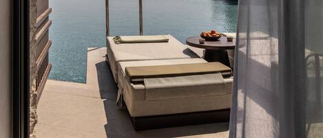 A balcony with a pool, two lounge chairs, a table with fruits, and a view of the sea.