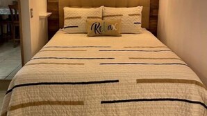 Quarto com cabeceira de madeira, duas luminárias de cabeceira, cama com colcha bege e travesseiro com a palavra 'Relax'.