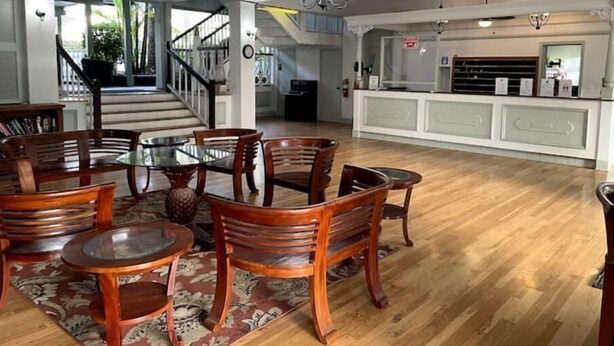 Uma sala de estar ampla com piso de madeira, uma mesa de jantar com tampo de vidro rodeada por cadeiras, uma escada que leva a um andar superior e uma cozinha com bancada branca e eletrodomésticos.