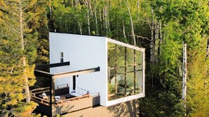 A modern cabin with a large glass window, wooden deck, and surrounded by dense forest. - World famous architect Design + Build (Tofte)