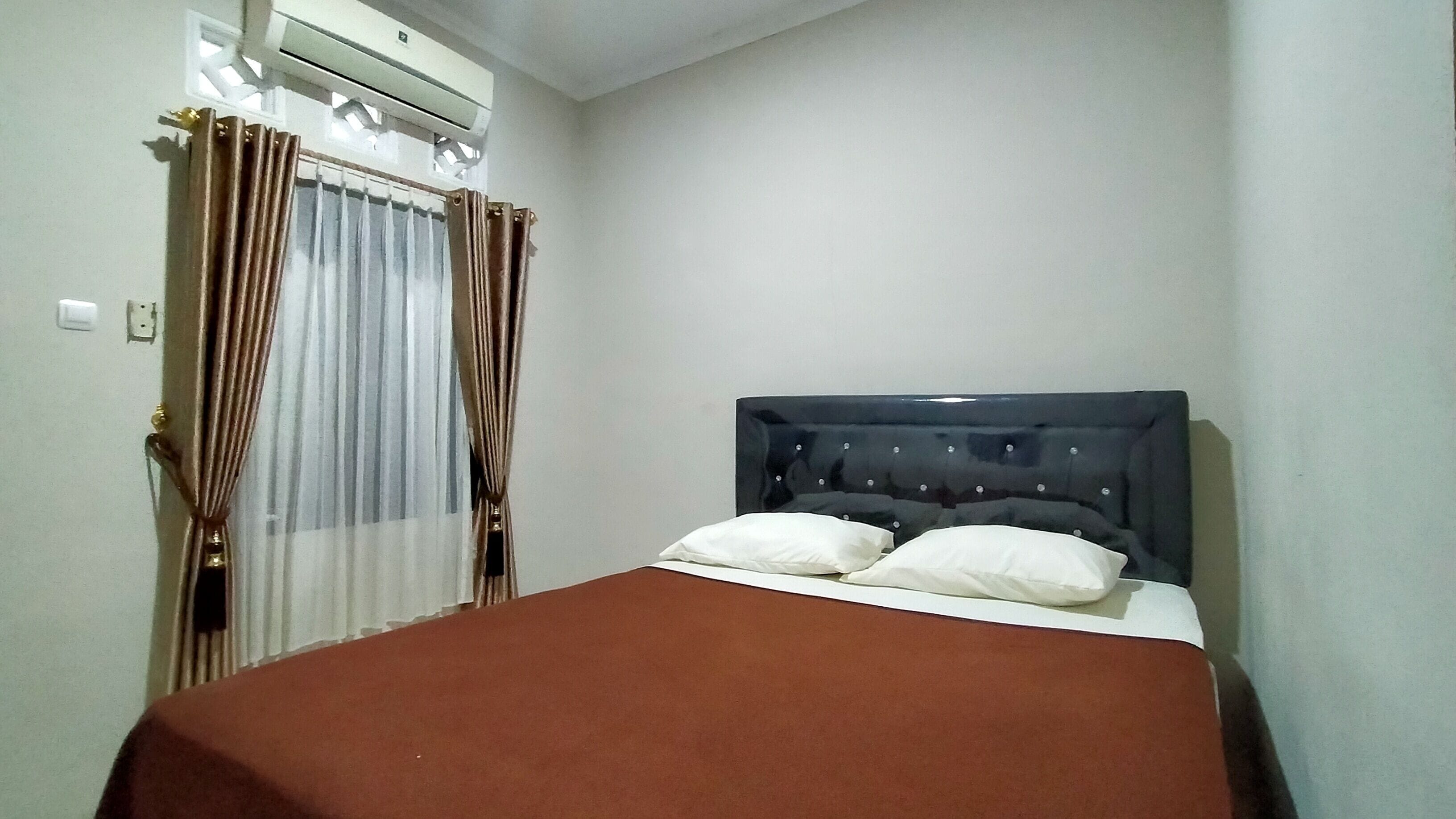 A single bed with a dark headboard and two pillows, a brown bedspread, and a window with curtains.