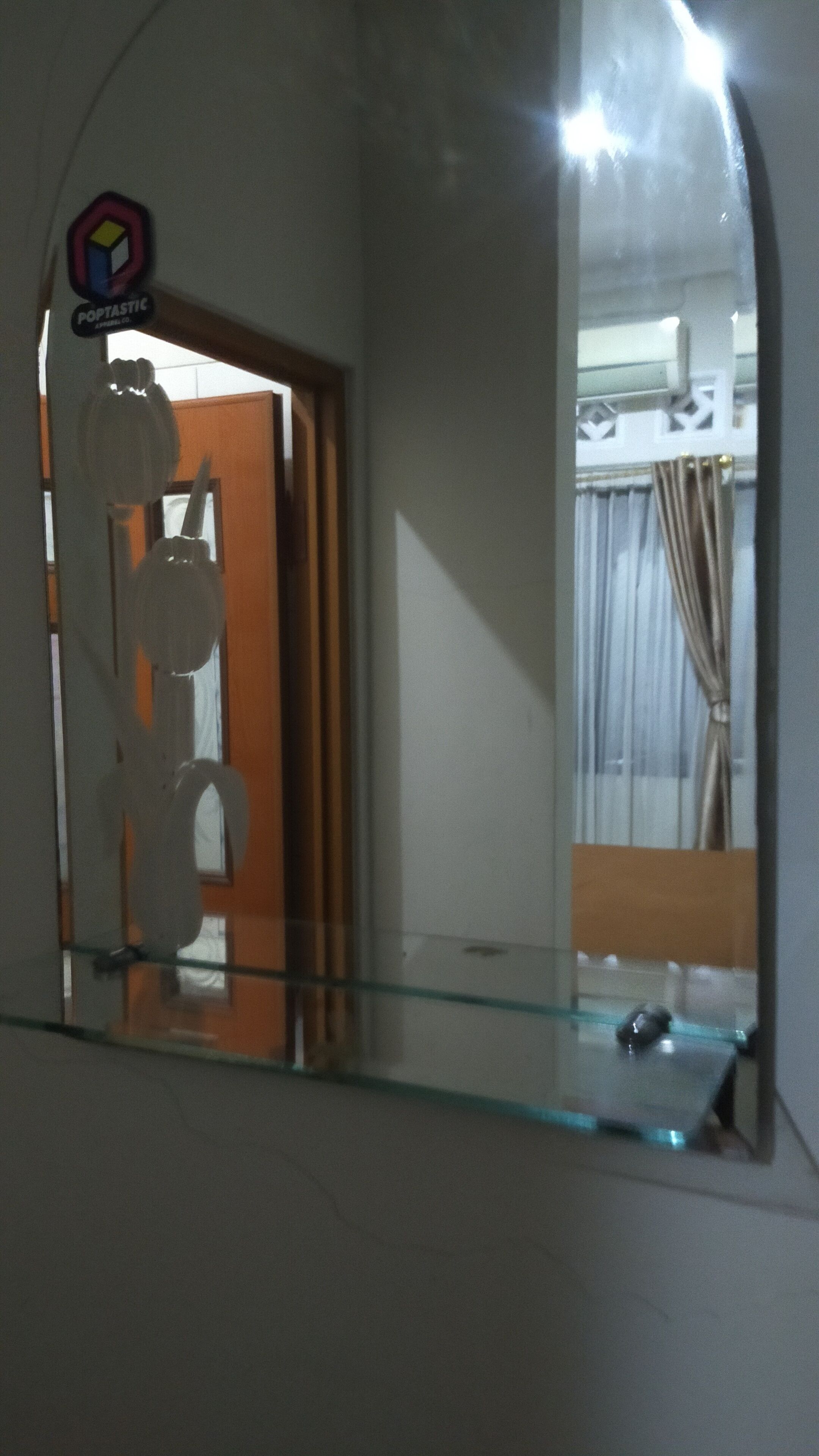 A bathroom with a glass shower enclosure, a visible door, and a reflection of a room with a bed and curtains.