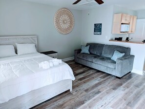 A bedroom with a bed, a grey sofa, a wooden floor, and a kitchen area with cabinets. - Cozy Studio Apt in Downtown MB (Myrtle Beach)
