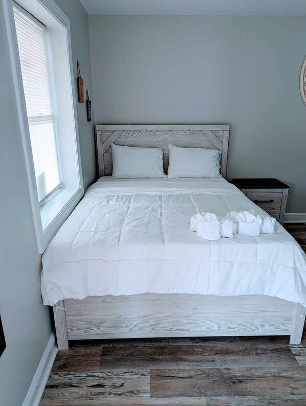 A neatly made bed with white linens and pillows, a wooden headboard, a nightstand with a drawer, and a window with blinds. - Cozy Studio Apt in Downtown MB (Myrtle Beach)