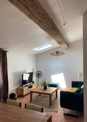 Ein Wohnzimmer mit Holzbalken, einem Fernseher auf einem Holztisch, einem Ventilator, einem Couchtisch mit Pflanze und einem grünen Sofa.