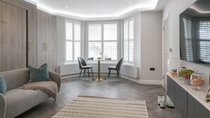 A modern living room with a sofa, a round table, and a bay window with white shutters.