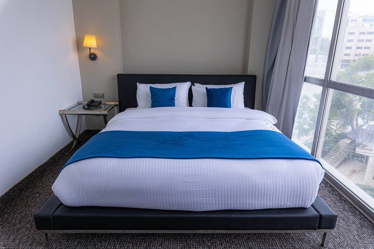 A hotel room with a large bed, a bedside table with a lamp, and a view of the city through the window.