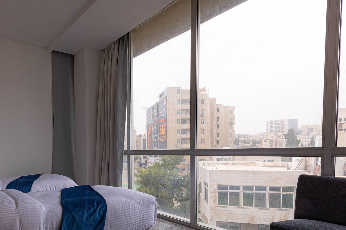 A hotel room with a bed, a lamp, and a large window offering a view of the cityscape.
