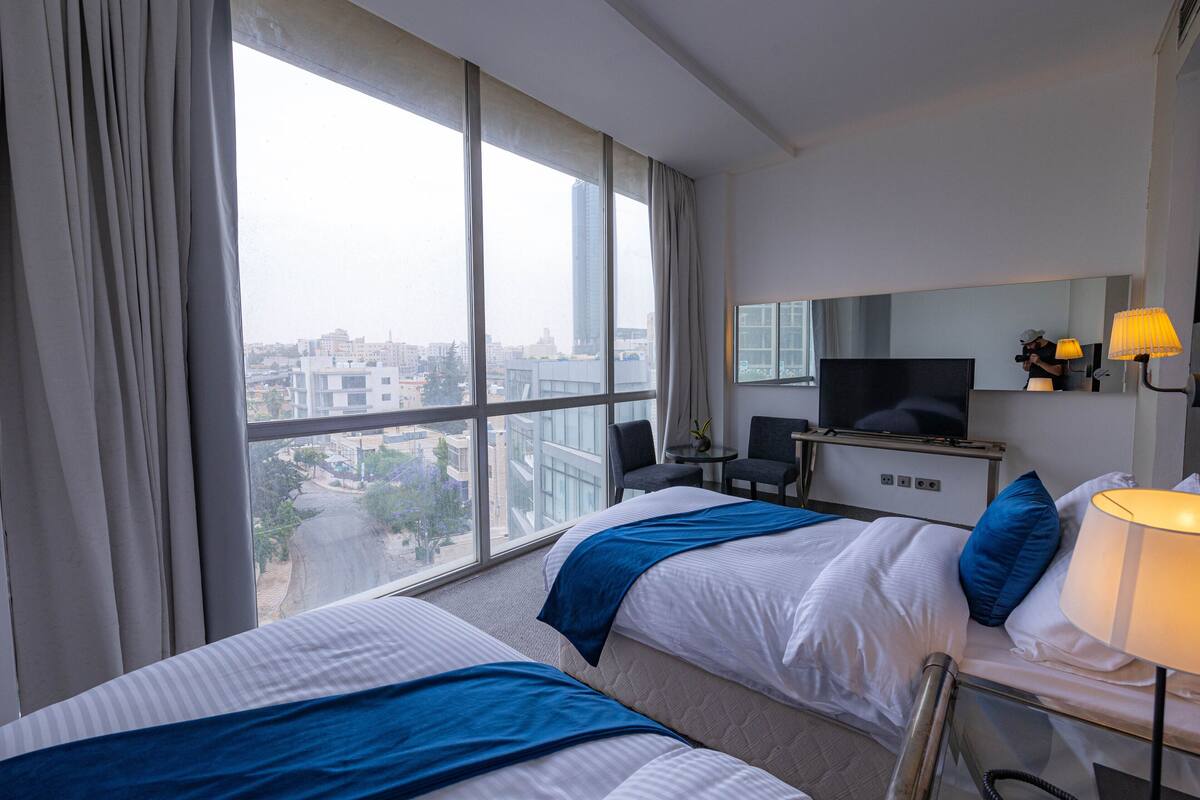 A hotel room with two beds, a TV, a desk with a lamp, and a view of the city.