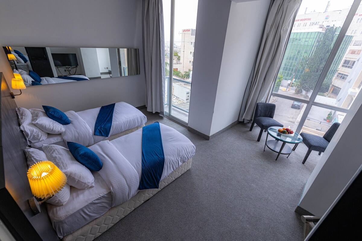 A hotel room with a large bed, a small table with chairs, a TV, and a view of the city through large windows.