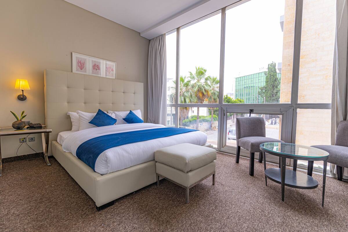 A hotel room with a large bed, a glass table, a chair, and a view of palm trees through the window.