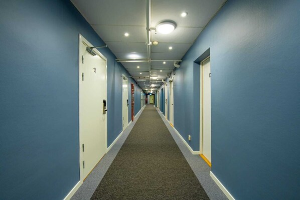 Un long couloir étroit, typique des hôtels, avec des murs bleus, un sol recouvert de moquette et des portes numérotées de 1 à 12.