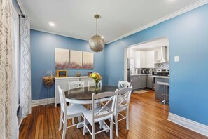 A dining area with a round table, white chairs, a hanging light fixture, and a kitchen in the background.