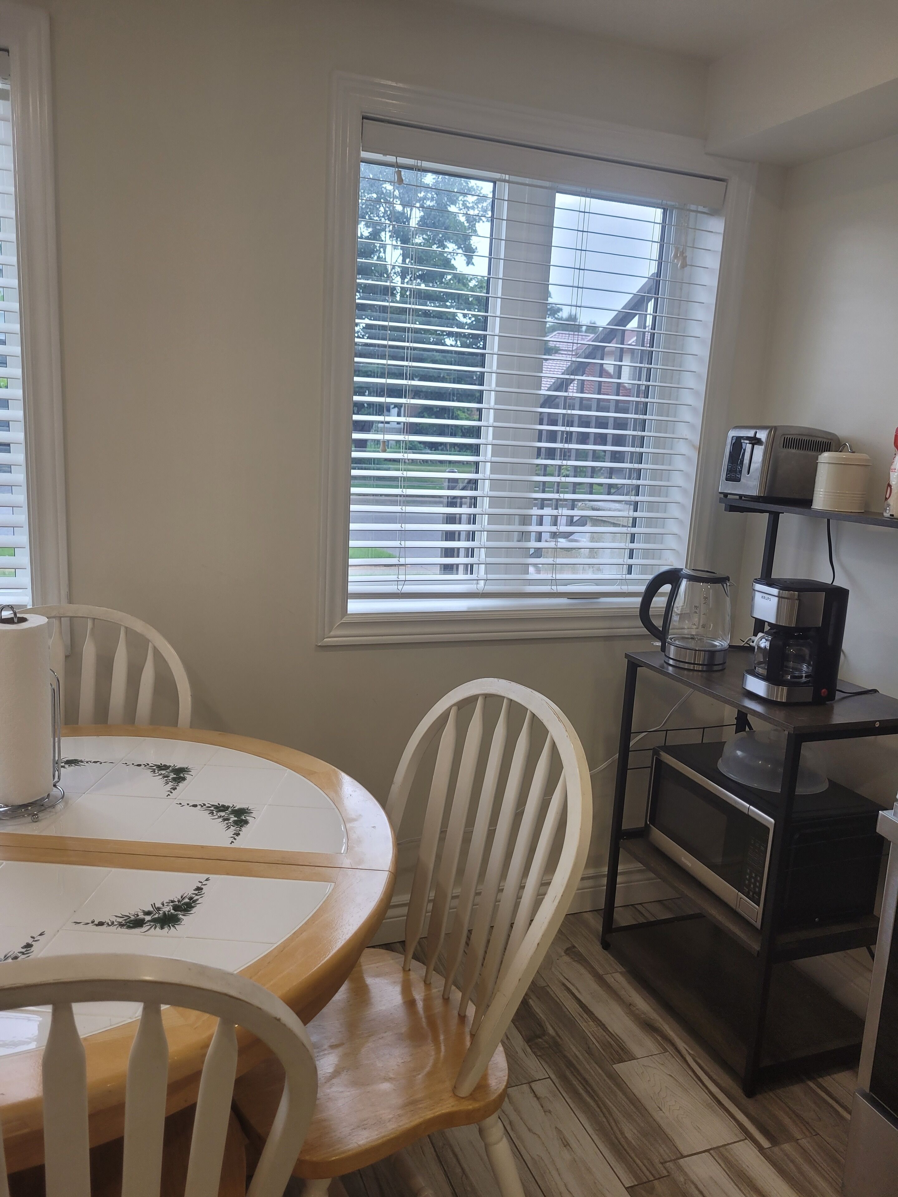 A small kitchen with a dining table, chairs, a coffee maker, a microwave, and a window with blinds.