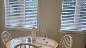 A dining area with a round table and four white chairs, two windows with blinds, and a wooden floor. - Cozy and modern bachelor suite with kitchenette and bath  (Guelph)