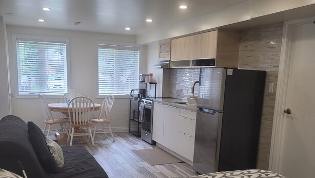 A modern, compact living space with a kitchenette, a dining area, and a bedroom with patterned bedding. - Cozy and modern bachelor suite with kitchenette and bath  (Guelph)