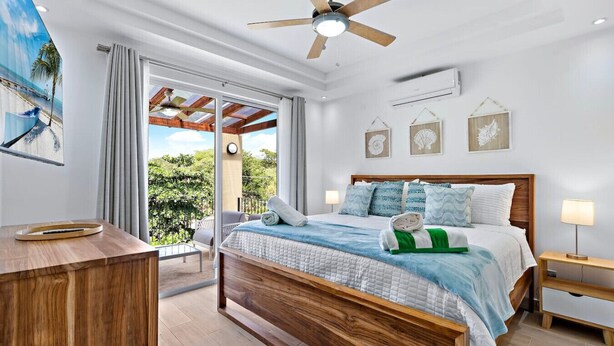 A bedroom with a wooden bed, a ceiling fan, and a view of greenery outside. - Gav5: New Modern Condo Walking Distance to Beach (Tamarindo)