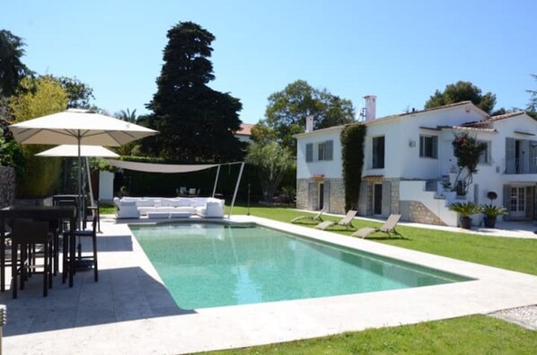 A rectangular swimming pool with a white deck, surrounded by a grassy lawn and a white house with a stone facade. - Charming villa close to Beach and old Antibes. (Antibes)