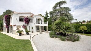 Una casa a due piani con facciata bianca, un balcone e un giardino curato con diverse piante e una palma.