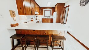 Una cocina con encimera blanca, armarios de madera y un lavamanos integrado. Hay tres taburetes debajo de la encimera.
