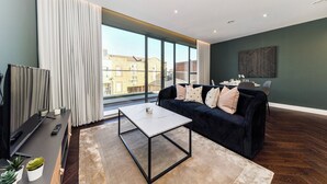 A modern living room with a dark green wall, a black sofa with patterned cushions, a white coffee table, and a large window with curtains. - Portobello Rd - North Ken by Viridian Apartments (London)