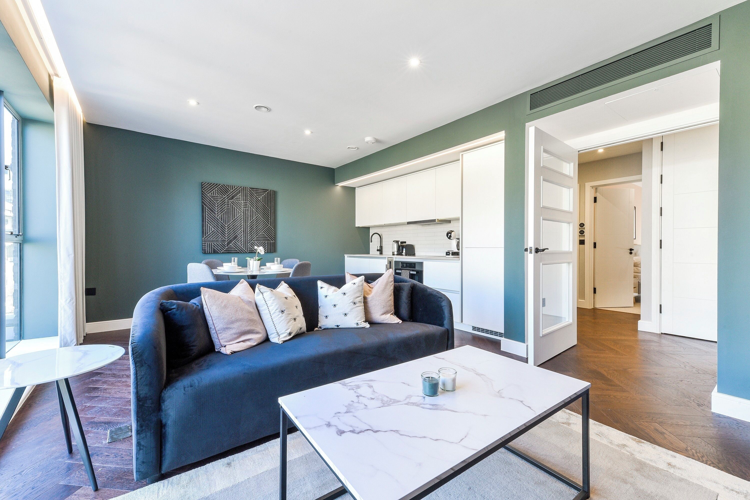 A modern living room with a dark blue sofa, a white coffee table, and a kitchen in the background.