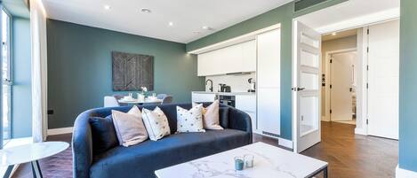 A modern living room with a dark blue sofa, a white coffee table, and a kitchen in the background.