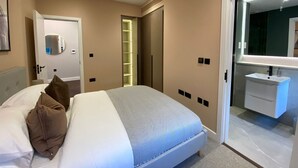 A modern bedroom with a bed, a bedside mirror, a walk-in closet, and a bathroom with a sink and mirror. - Portobello Rd - North Ken by Viridian Apartments (London)