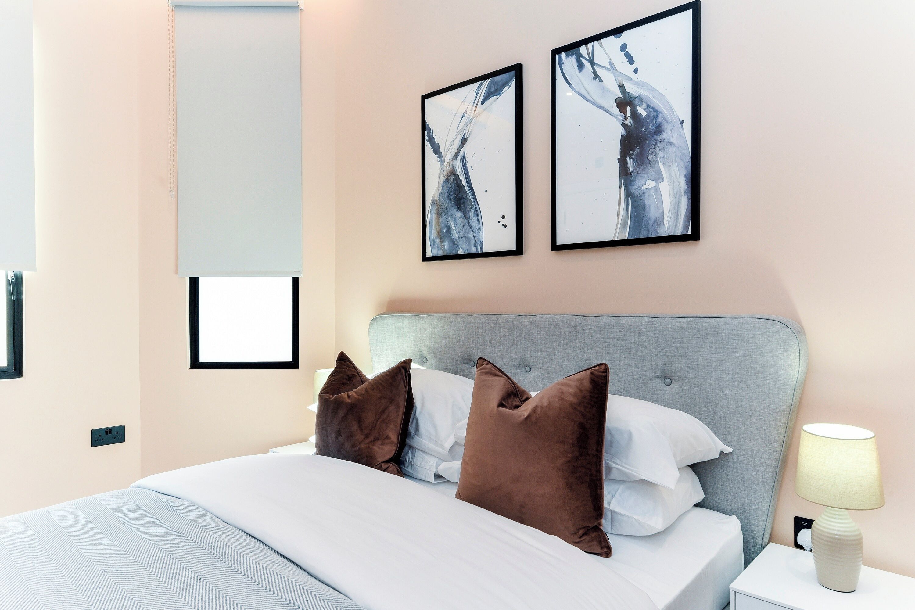 Un lit bien fait, avec une tête de lit grise, deux oreillers marron et une couette blanche. Deux tableaux abstraits encadrés sont accrochés au mur au-dessus du lit.