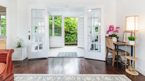 Una habitación con suelo de madera, paredes blancas, una silla de madera, una pequeña mesa con una lámpara y vistas al exterior con vegetación.