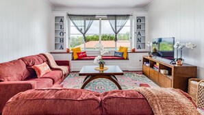 Una sala de estar acogedora con un sofá rojo, una mesa de centro de madera y una estantería. Se ve una ventana con cortinas y vistas al verdor exterior.