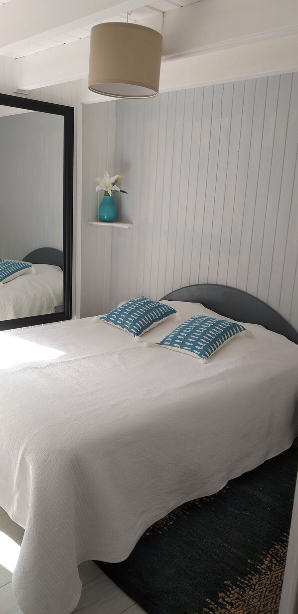 Un lit bien fait, avec une literie blanche et des coussins à motifs bleus, un petit vase turquoise contenant une fleur blanche, posé sur une étagère blanche, et un grand miroir accroché au mur.