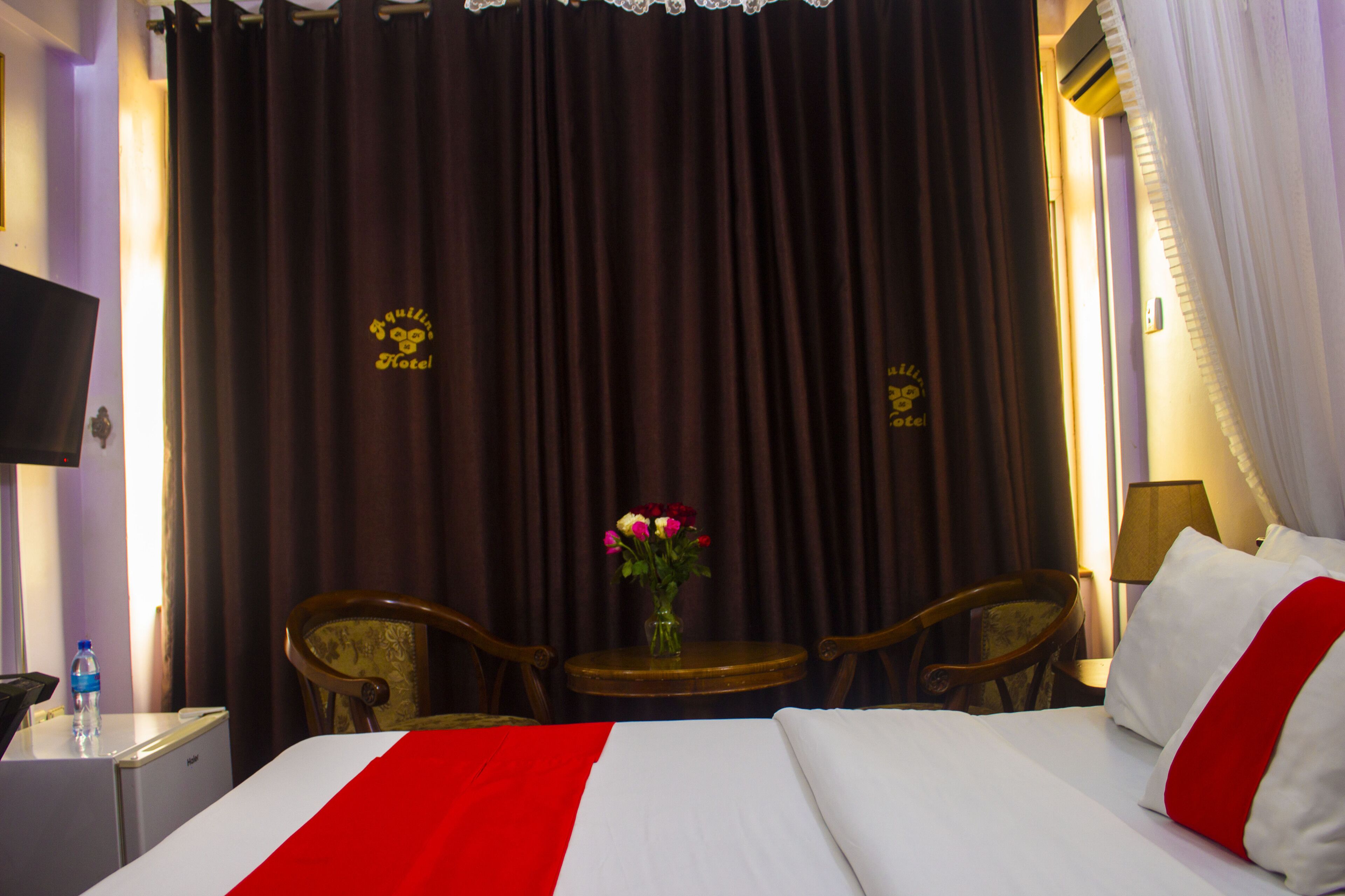 Habitación de hotel con cama, una mesita con un florero, dos sillas y un televisor sobre una mesita de noche.