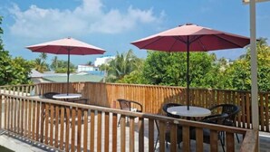 Een terras buiten met houten hekwerk, twee ronde tafels en twee rode parasols.
