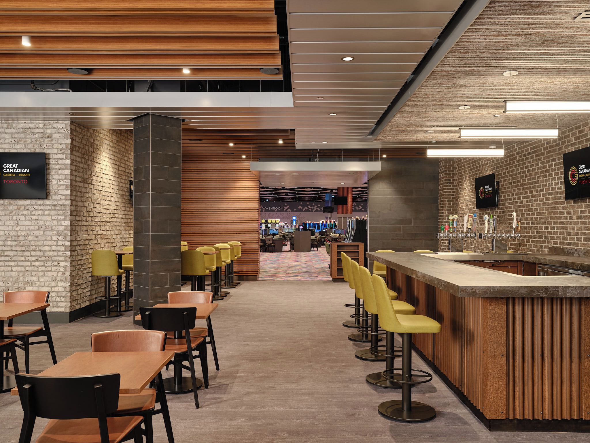 A modern bar area with a long counter and multiple stools, a seating area with tables and chairs, and a visible washroom in the background.