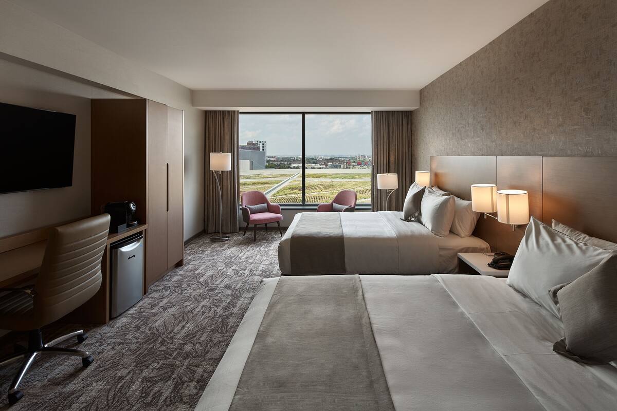 A hotel room with a large bed, a desk, a chair, and a TV. There is a window with a view of the city.
