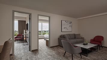 A modern hotel room with a grey sofa, a red armchair, a grey armchair, a small table with a plant, and a large window offering a view of the city.