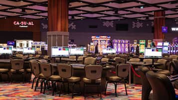 Un salón de casino con varias mesas de juego y sillas, una alfombra colorida y máquinas tragamonedas.