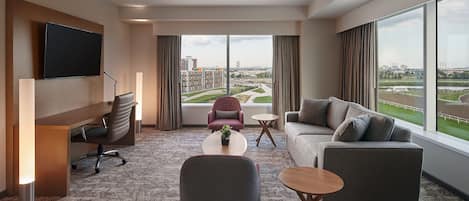 A modern hotel room with a large window, a flat-screen TV, a desk, a sofa, and a coffee table.