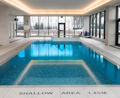 A swimming pool with a shallow area marked, surrounded by a modern indoor setting with large windows.