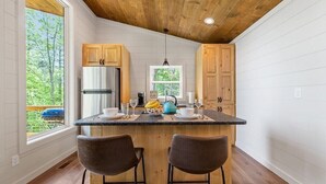 A modern kitchen with wooden cabinets, a central island with bar stools, a refrigerator, and a window with a view of greenery. - Dream Big in this new Tiny Home! Hot tub, firepit (Mineral Bluff)